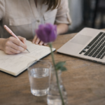 Woman writing in notebook beside laptop while planning automated webinar funnel