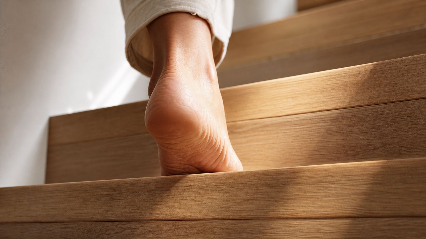 A close-up view of a bare human foot stepping up on wooden stairs at home.